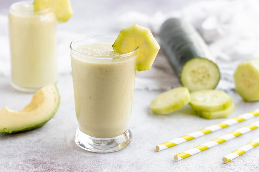 a green cucumber smoothie sits in front of a sliced cucumber, avocado and straws
