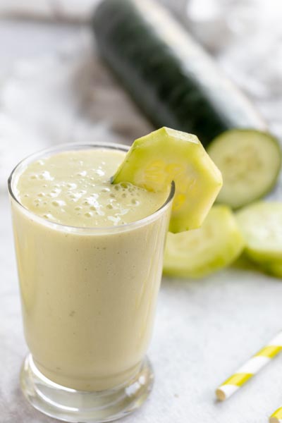 a creamy green smoothie in a glass with a large cucumber in the background
