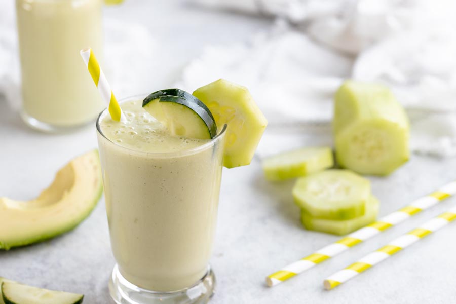 a glass with a cucumber smoothie in it and a few slices of cucumber with straws and avocado near