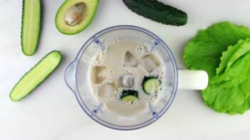 ice, cucumber and other ingredients in a blender with a slice of avocado near by