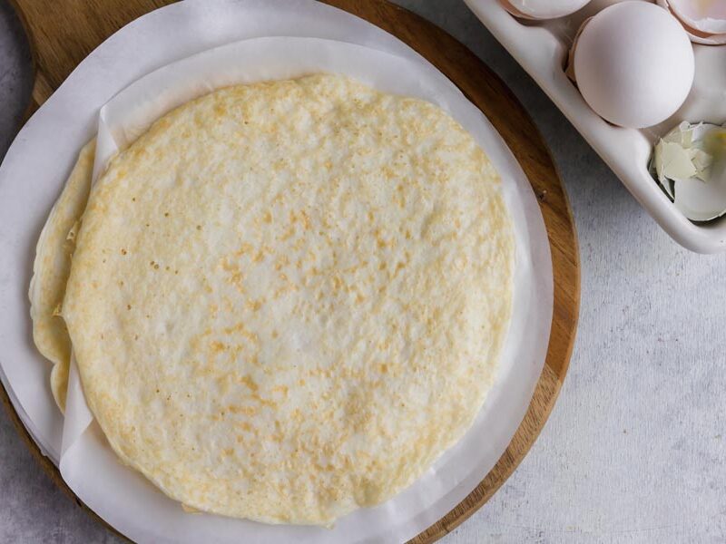 Looking down on a stack of egg wraps on a cutting board next to a carton of eggs.