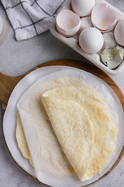 Multiple egg wraps on a cutting board next to a 6 pack of eggs with whole eggs and egg shells.