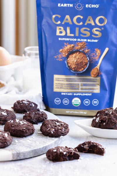 a blue bag of Cacao Bliss sits behind chocolate cookies on a counter with a glass of milk in the background