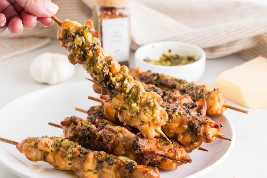 A handing holding a chicken skewer over a plate. Each skewer is coated with a garlic parmesan parsley sauce.