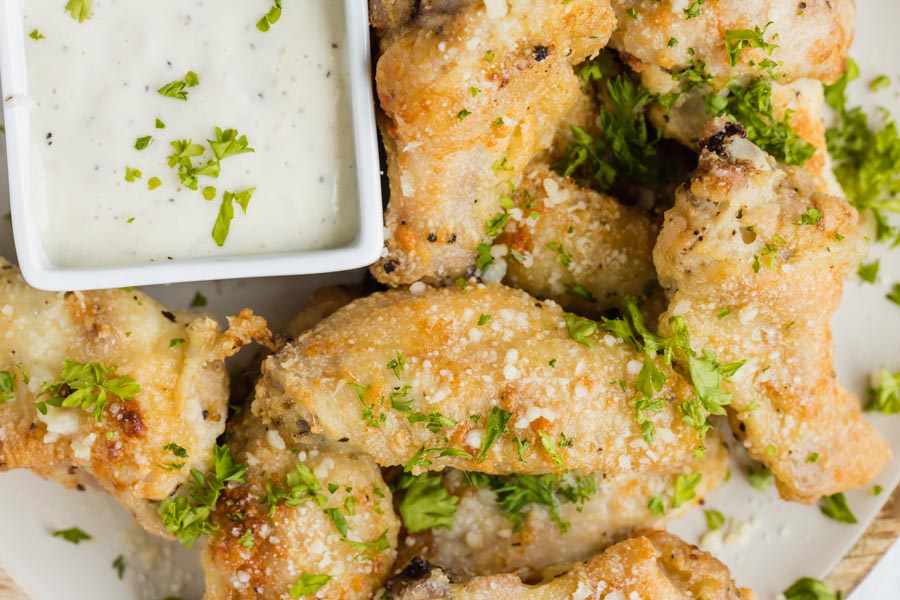 Looking down onto a plate of crispy party wings cooked golden brown and coated with grated parmesan and minced parsley. They sit next to a small bowl of ranch.