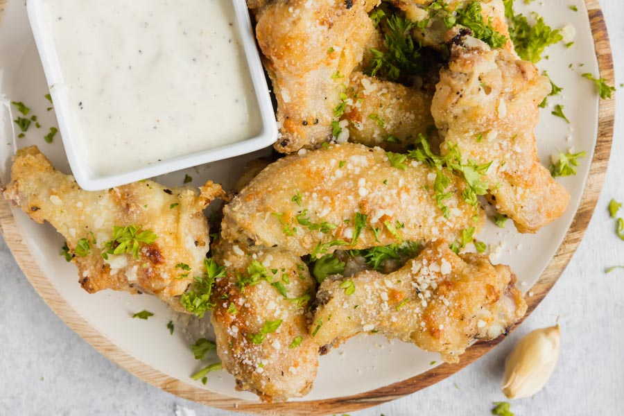 A plate filled with party wings coated with minced parsley and grated parmesan cheese next to garlic cloves and ranch dressing.