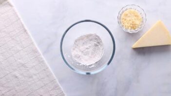 A small clear bowl with a white baking powder mixture inside. A slice parmesan sits nearby.