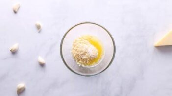 A small bowl with melted butter, minced garlic and shredded parmesan next to cloves of garlic.