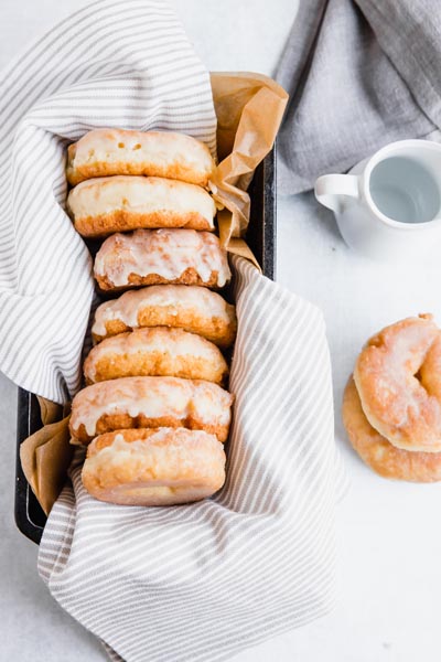 glazed donuts in a loaf pan lined with a towel next to two donuts stacked and a creamer jar