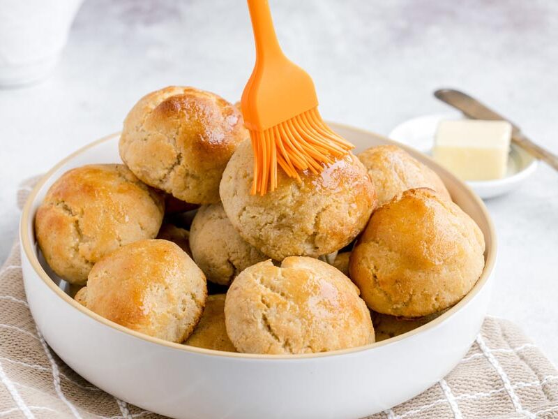 A batch of golden keto yeast rolls being brushed with melted butter in a white bowl.
