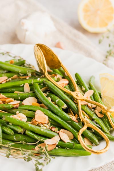 toasted almond slices mixed with sauteed green beans next to gold tongs and fresh thyme