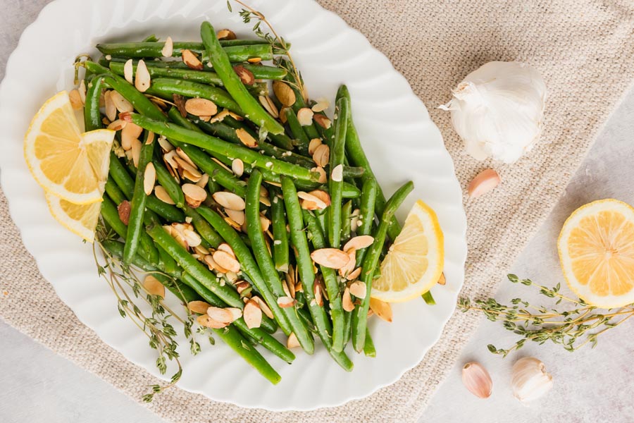 cooked green beans on a plate with three slices of lemon and toasted almond scattered over the beans