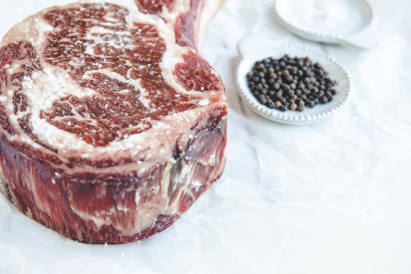 thick up of ribeye steak with lots of marbling next to two small plates with salt and peppercorns