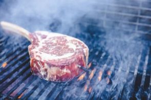 tomahawk steak on a grill over the flames with a meat thermometer stuck inside the meat