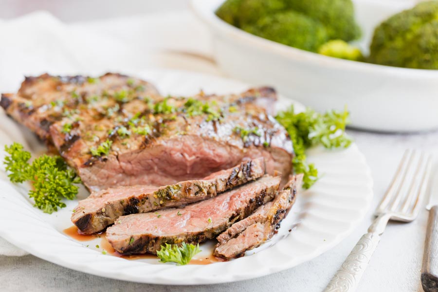 Juicy flank steak on a platter with a few pieces sliced and juices coming out. Steak is cooked medium rare. A dish of broccoli nearby.