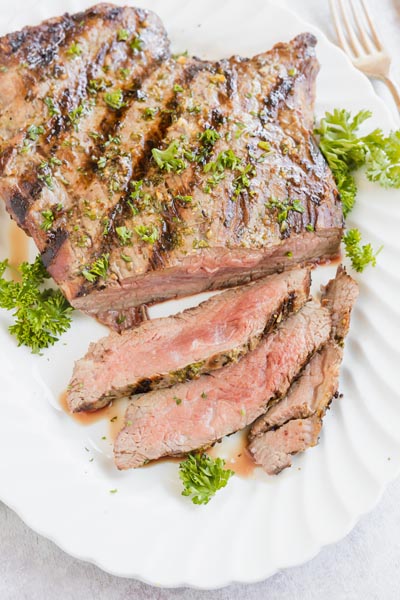 A platter with sliced flank steak cooked medium rare on it. Half of the steak is sliced and other other is whole. A fork and parsley are nearby.