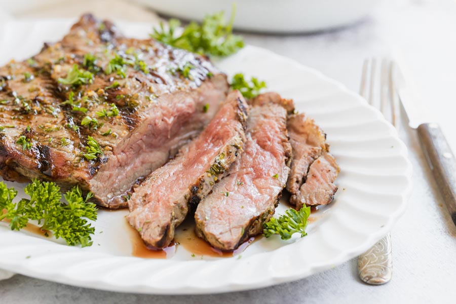 Medium rare slices of steak spilling juices on a white platter and covered with chopped parsley.