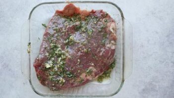 A large chunk of beef marinating in a herb garlic marinade in a baking dish.