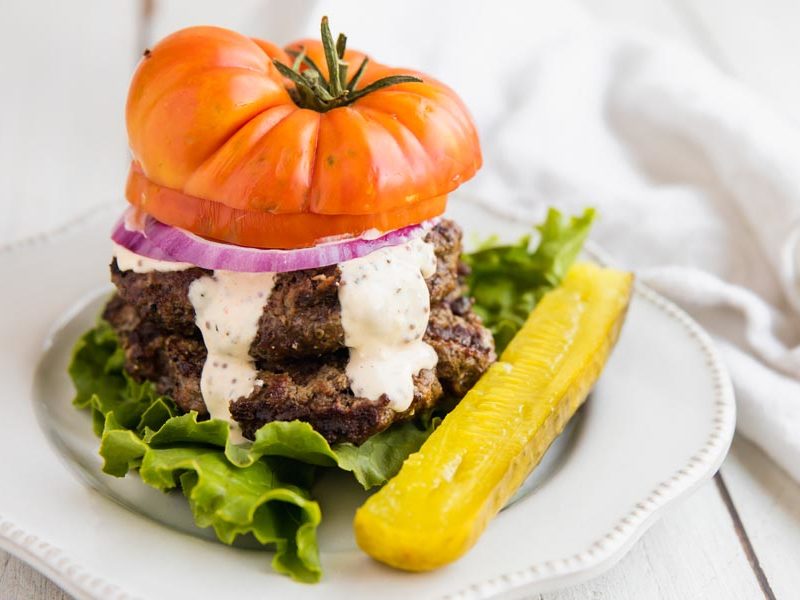 a bunless burger with a tomato slice on top and lettuce on the bottom with a side of pickle spear