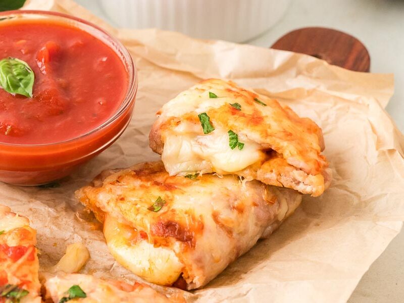 Cheesy chicken calzones stacked beside a bowl of marinara sauce for dipping.
