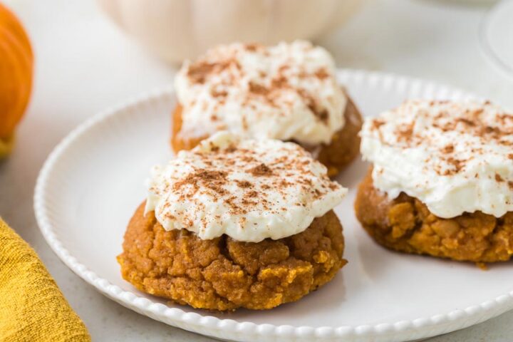 Three frosted pumpkin cookies topped with cinnamon sit on a white scalloped plate.
