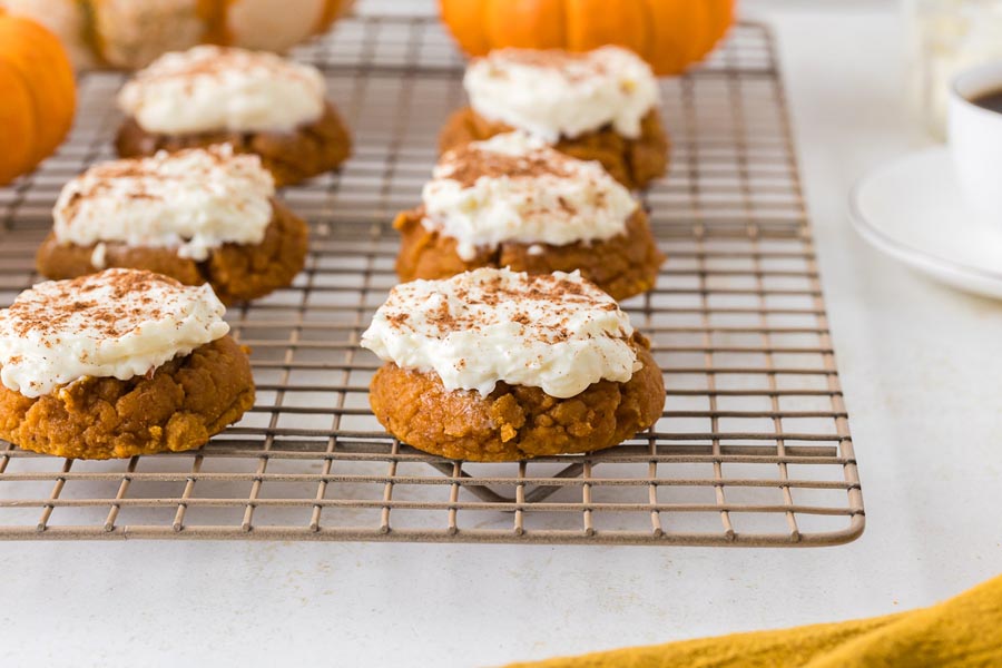 Pumpkin cookies with white frosting and a dusting of cinnamon cool on a wire rack surrounded by mini pumpkins.