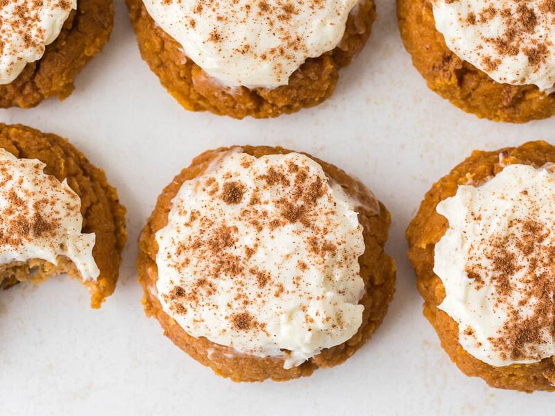 Overhead view of pumpkin cookies with cream cheese frosting and cinnamon, one showing a bite taken out.