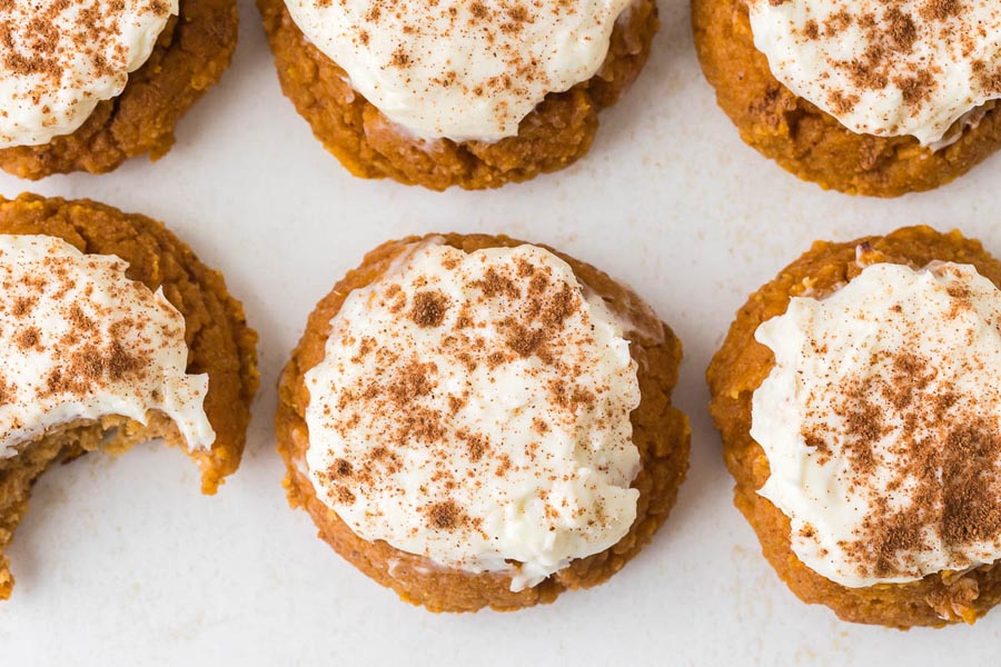Overhead view of pumpkin cookies with cream cheese frosting and cinnamon, one showing a bite taken out.