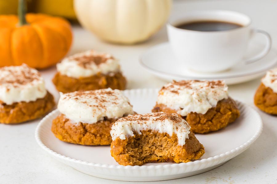 A bitten pumpkin cookie sits on a plate next to others, with coffee and pumpkins in the background.