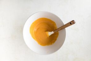A white mixing bowl holds a smooth orange pumpkin mixture with a spatula.