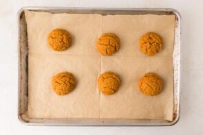 Six plain pumpkin cookies are spaced out on parchment paper on a baking tray.