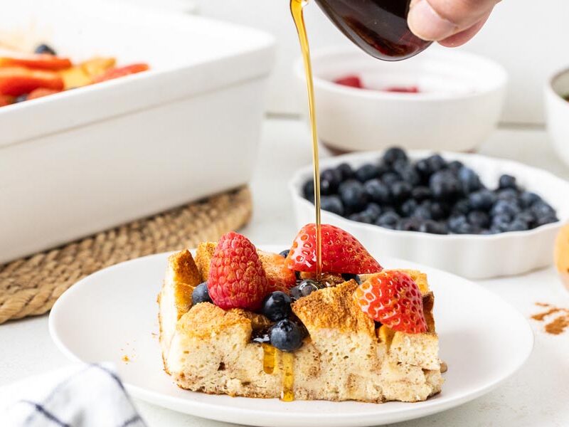 A hand pouring syrup over a slice of french toast casserole that is topped with raspberries, blueberries and strawberry slices.