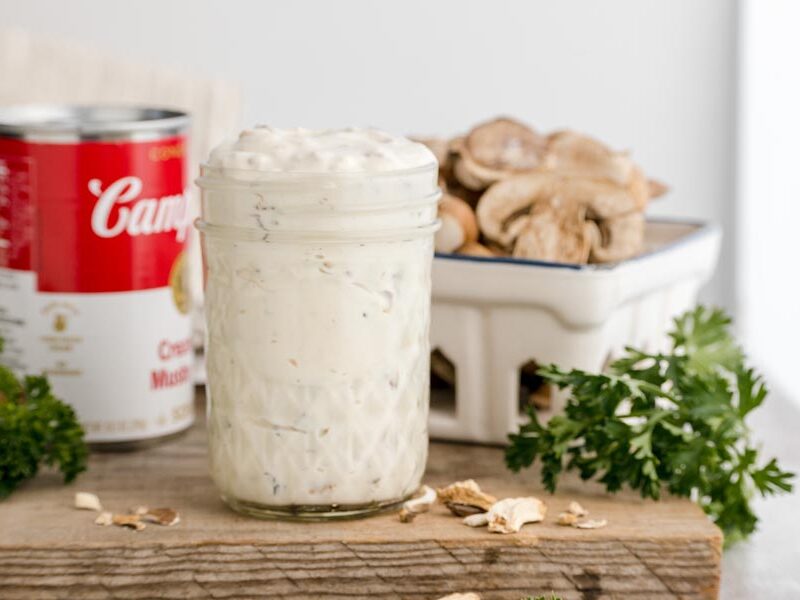 Keto cream of mushroom soup in a glass jar displayed beside fresh mushrooms and a can of condensed mushroom soup.