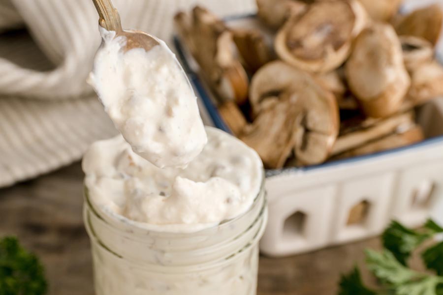A spoonful of thick homemade cream of mushroom soup lifted from a glass jar with fresh mushrooms in the background.