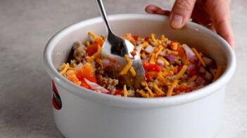 Cooked sausage, shredded cheese, peppers, onions, and seasonings being mixed in a round cake pan.