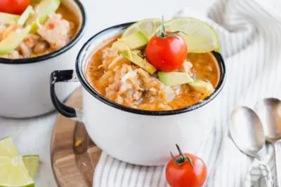 hot mug of chicken and cauliflower rice topped with a cherry tomato and limes with two spoons next to it