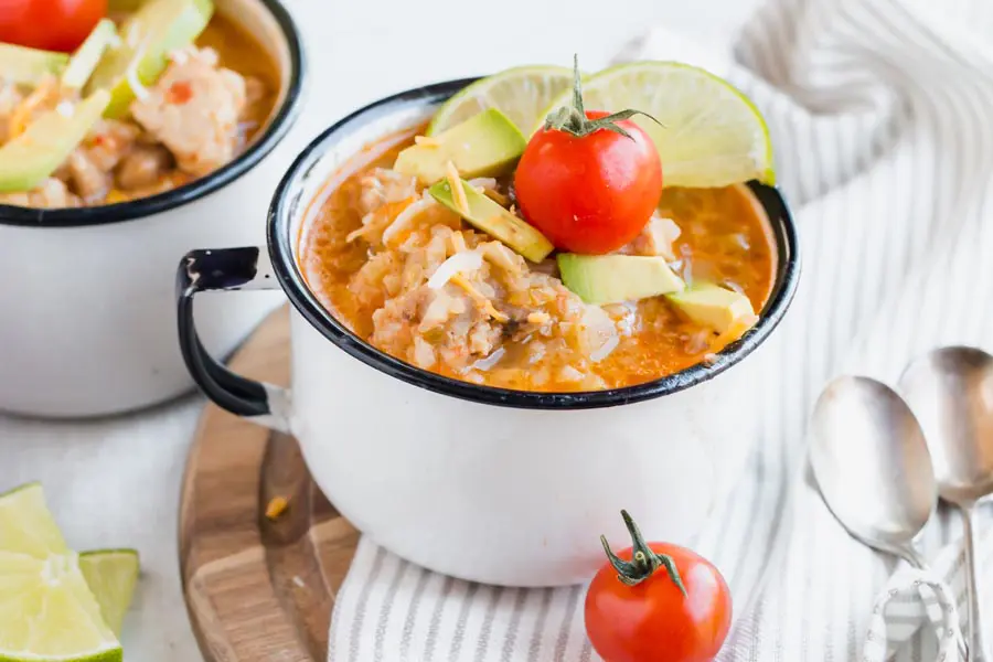 hot mug of chicken and cauliflower rice topped with a cherry tomato and limes with two spoons next to it