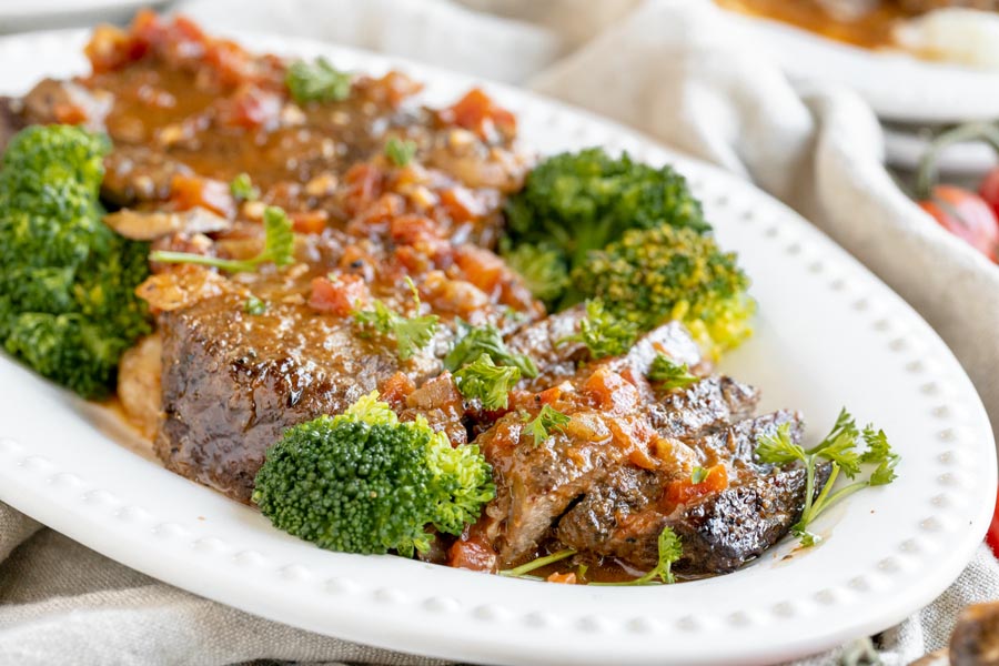 juicy slices of steak cu on a platter topped with a tomato liquid and broccoli and parsley around