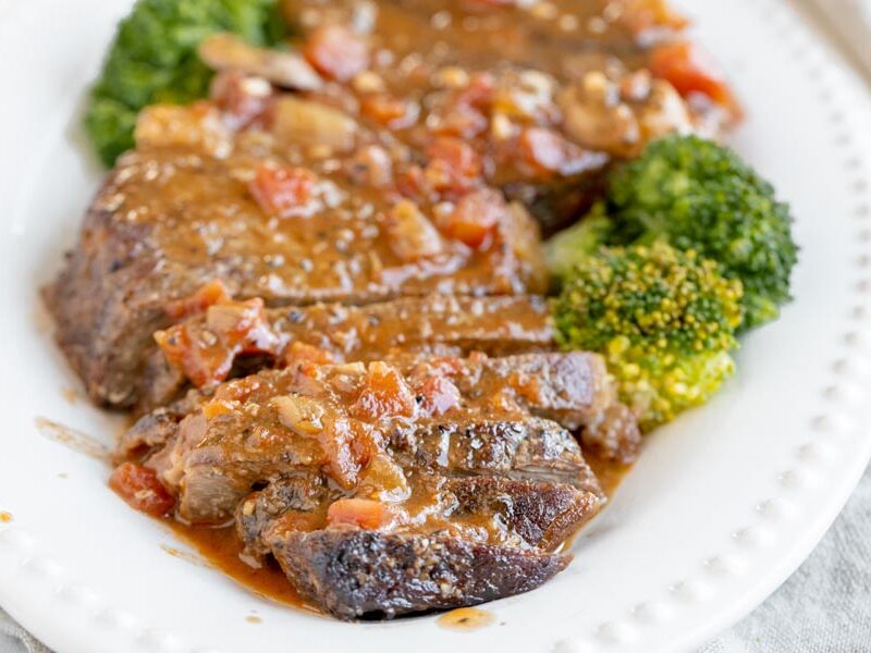 juicy steak sliced on a platter and covered in a tomato based sauce with broccoli florets on the platter too