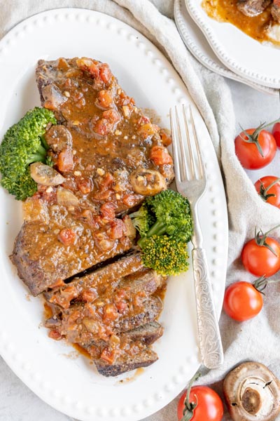 a platter with swiss steak on the dinner table with broccoli and tomatoes around