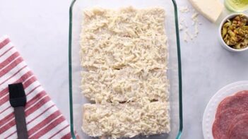 Shredded white cheese on top of the bottom halves of dinner rolls in a clear baking dish.