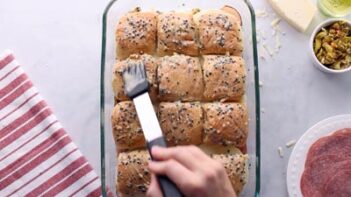 A hand brushes a buttery mixture of onion and black sesame seeds on top of sandwich rolls.