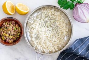 A skillet with prepared cauliflower rice sits next to a sliced red onion, lemon, parsley and nuts.