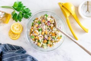 A salad in a glass bowl with a wooden spoon stuck in the salad. A lemon squeezer sits nearby.