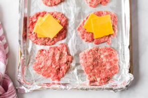 A tray with burger patties. Some have slices of cheese on them.