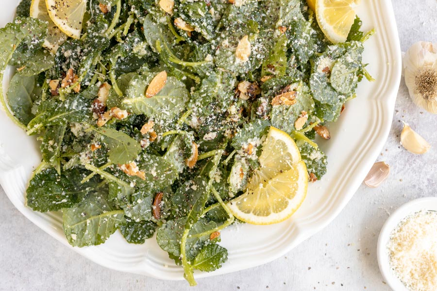 A large platter of tossed kale salad with lemon slices, grated parmesan chees and garlic cloves nearby.