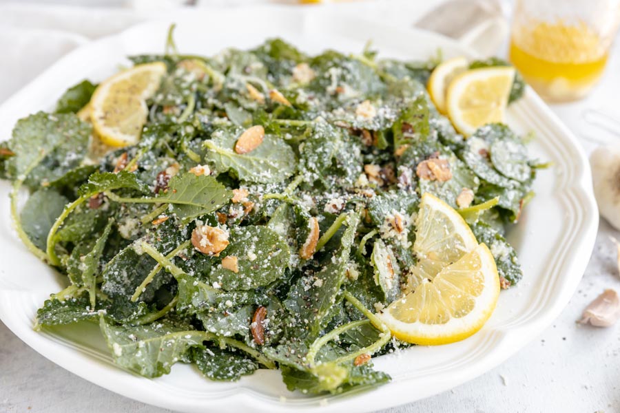 Toss fresh kale salad from baby kale leaves, toasted almond slices and parmesan cheese on a plate with lemon slices.