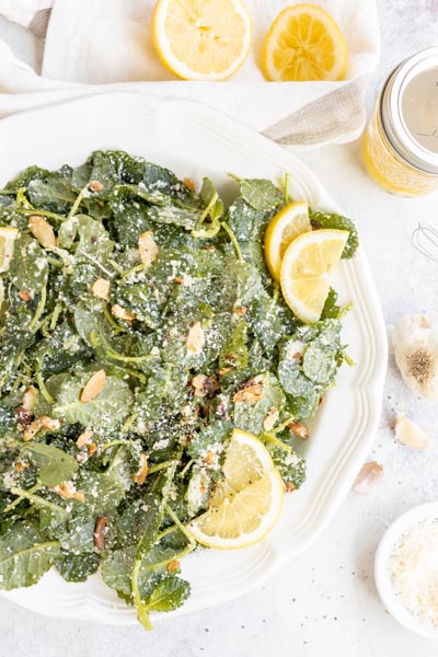 Looking down onto a platter with fresh lemon garlic kale salad topped with almonds and parmesan cheese by freshly squeezed lemons, garlic and dressing.