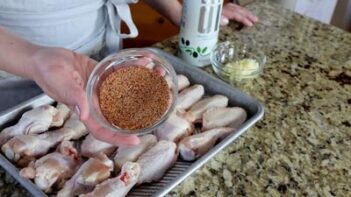 Annie from KetoFocus holding a bowl of seasoning over the chicken wings lined in a baking sheet.