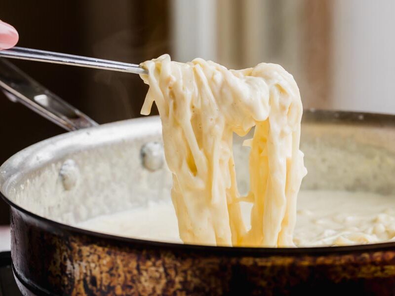 Holding a spoonful of creamy fettucine alfredo over a skillet.